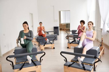Bir grup sportif kadın bir odada pilates yapıyor, sandalyede oturuyor..