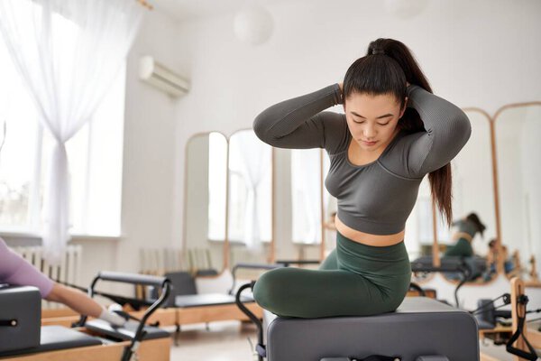 Asian woman in gray top and green pants works out in gym, next to her friend.