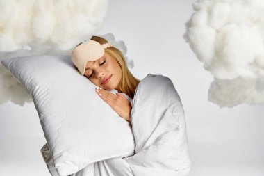 Blonde woman in pyjamas lies on cloud pillow with serene expression. clipart