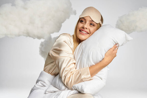 Dreamy blonde woman in cozy pajamas sitting amongst clouds with a pillow.