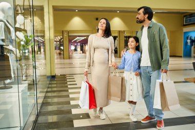 Mutlu bir aile kalabalık bir alışveriş merkezinde yürüyor, alışveriş torbaları taşıyor ve haftasonu gezintisinin tadını çıkarıyor..
