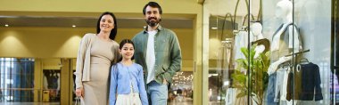 Neşeli bir aile, ebeveynler ve çocuklar, hareketli bir alışveriş merkezinde hafta sonları gezintiye çıkan enerjik dükkanların arasında yürüyorlar..