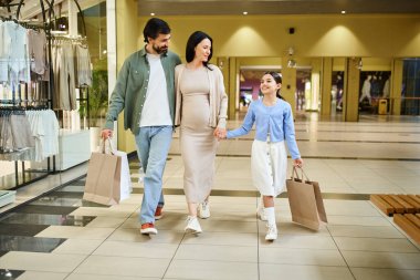 Alışveriş torbaları taşıyan mutlu bir aile, hafta sonu gezintisi için kalabalık bir alışveriş merkezinde rahatça dolaşır..