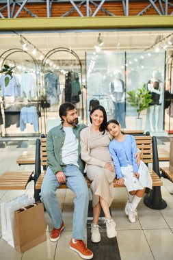Mutlu bir aile, alışveriş haftasonunun tadını çıkarıyor, kalabalık bir alışveriş merkezinde bir bankta birlikte oturuyor, beraberce bir anı paylaşıyor..