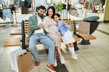 Mutlu bir aile kalabalık bir alışveriş merkezinde bir bankta oturup hafta sonu gezintisinin tadını çıkarıyor..
