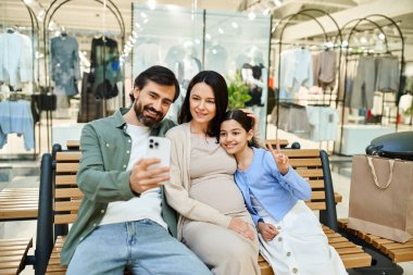 Neşeli bir aile alışveriş merkezindeki bankta bir anı yakalar, birlikte selfie çekerken gülümserler..