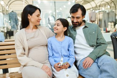 A pregnant woman and her daughter share a tender moment on a bench in a bustling shopping mall. clipart