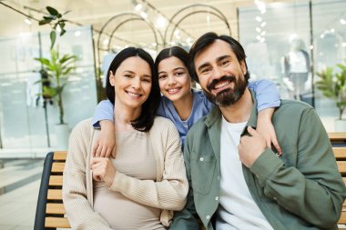 Neşeli bir aile, hareketli bir alışveriş merkezindeki bankta oturur ve hafta sonu gezileri sırasında paylaştıkları anın tadını çıkarırlar..