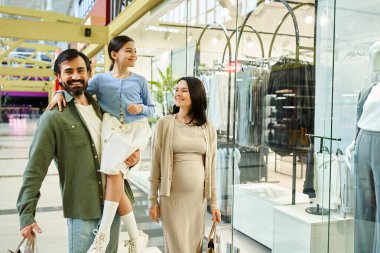 Mutlu bir aile, ebeveynler ve çocuklar, mağazalar, insanlar ve renkli vitrinlerle dolu canlı bir alışveriş merkezinden rahatça geçiyorlar..