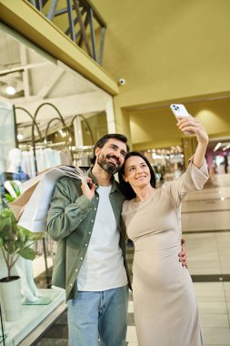 Hamile bir kadın ve erkek kalabalık bir alışveriş merkezinde alışveriş yaparken mutlu bir şekilde selfie çekiyorlar..