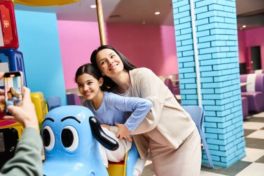 A mother and her daughter smile as they enjoy a ride on a toy carousel in a malls gaming zone on the weekend. clipart