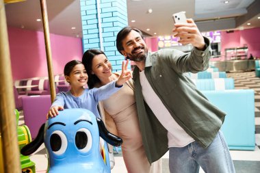 Neşeli bir aile hafta sonu alışveriş merkezlerinin oyun sahasında atlıkarınca oyuncağının önünde selfie çekerken gülümser..