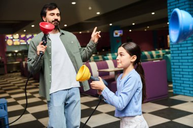 A father and his daughter happily play a game in a gaming zone within a mall on a weekend. clipart
