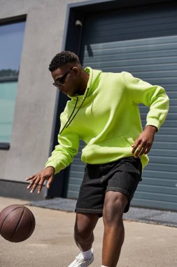 Yakışıklı Afrikalı Amerikalı adam neon kapüşonlu ve şort giyiyor, elinde basketbol topu var..