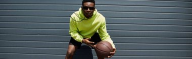 Yeşil kapüşonlu yakışıklı Afro-Amerikan adam elinde basketbol topu tutuyor..