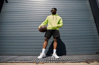 Young African American man in yellow hoodie playfully holds basketball. clipart