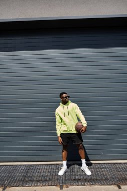 Yakışıklı, Afro-Amerikalı, şık giyinmiş, elinde basketbol topuyla bir garajın önünde..