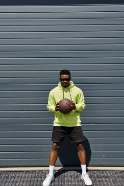 Havalı Afro-Amerikan bir adam elinde basketbol topuyla bir garajın önünde.