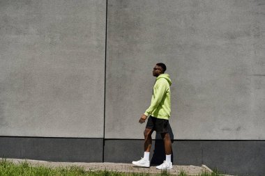 Neon yeşil kapüşonlu şık Afro-Amerikan bir adam duvarın yanından geçiyor..