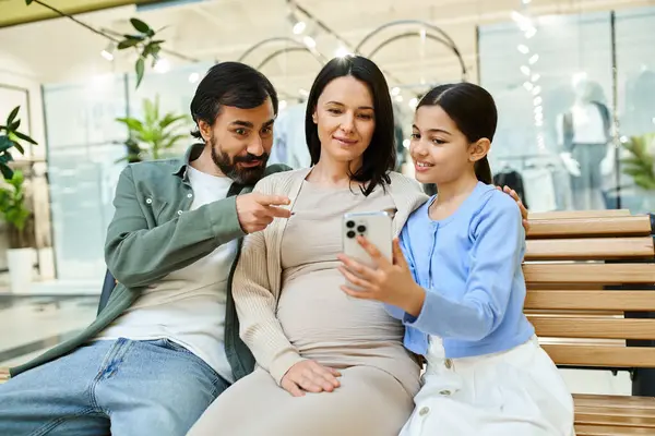 stock image A family sits closely on a bench in a shopping mall, engrossed in a cell phone screen together.