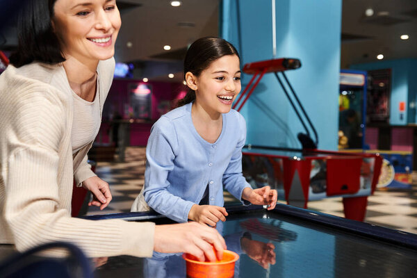 A joyful mother and daughter immerse themselves in a spirited air hockey match at a gaming zone in a mall on the weekend.