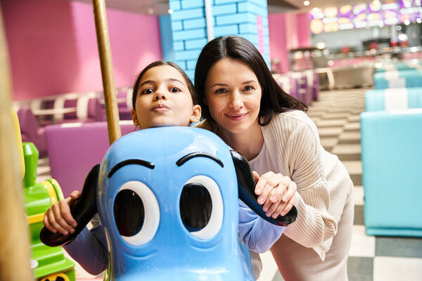 A cheerful woman and child joyfully posing together next to a colorful carousel toy in a mall gaming zone.