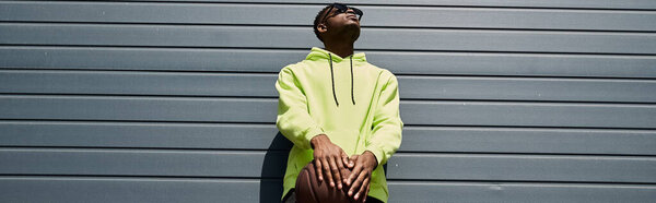 Красивый афроамериканец, прислонившийся к городской стене в стильной желтой толстовке.