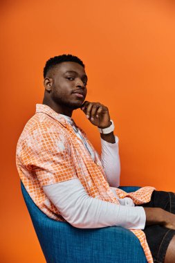 Parlak turuncu arka planlı, modern mavi sandalyede oturan, modaya uygun giyinmiş yakışıklı Afro-Amerikalı adam..