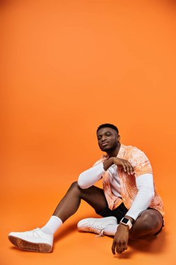 Parlak turuncu arka planda oturan Afrikalı Amerikalı şık bir adam..