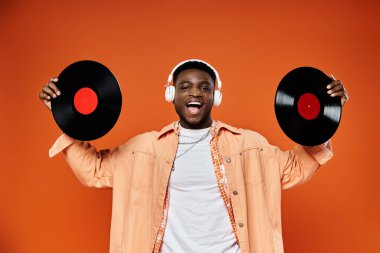 Stylish African American man holding vinyl records against orange backdrop. clipart
