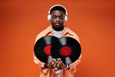 Handsome African American man in stylish attire holding a record and listening with headphones. clipart