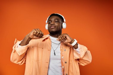 Stylish black man with headphones striking a pose against vibrant orange backdrop. clipart