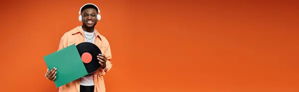 stock image Young African American man in fashionable attire holding a vinyl record in orange backdrop.