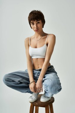 Young woman in white top, jeans, posing on stool.