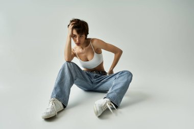 Young woman in jeans and white tank top sits on the floor in a stylish pose.