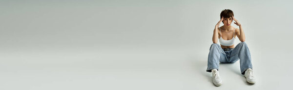 A young woman in stylish attire sits on the floor with her hands on her head.