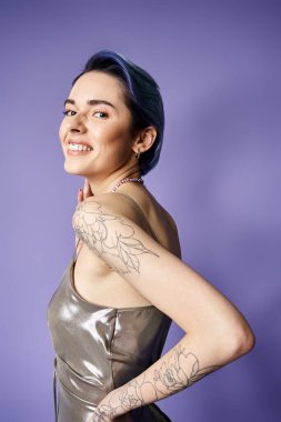 Young woman with short blue hair proudly displays a detailed tattoo on her arm in a silver party dress.