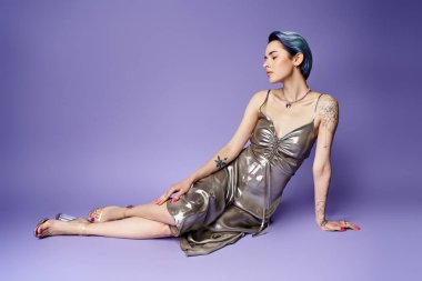 Young woman with short blue hair, in silver party dress, sitting gracefully on the floor in a studio setting.