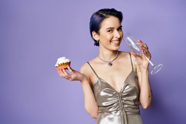 A stylish young woman with short blue hair in a silver dress holds a cupcake and a wine glass in a posed studio shot.