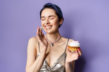 Young woman with short blue hair in a silver party dress holding a vibrant cupcake, exuding elegance and charm.