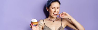 A pretty young woman with short blue hair in a silver party dress joyfully holds and savors a delicious cupcake.