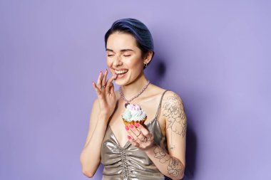 A stylish young woman with blue hair holds a beautifully decorated cupcake in a dazzling silver dress.