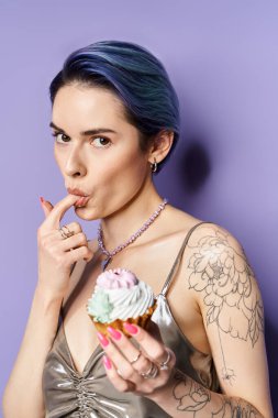A stylish young woman with short blue hair in a silver party dress holding a delicious cupcake.
