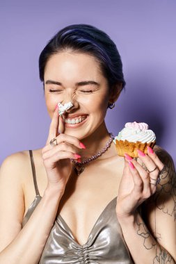 Young woman with short blue hair holding cupcake in front of her face, dressed in silver party attire, striking a whimsical pose.