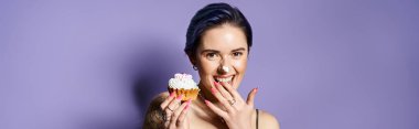 A pretty young woman with short blue hair in a silver party dress hides her face behind a delicious cupcake.