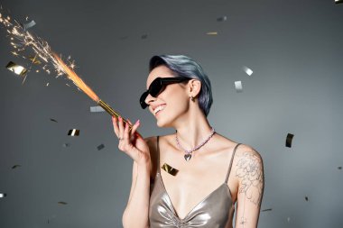 Young woman with short blue hair elegantly holds a sparkler, dressed in a glamorous silver outfit in a studio setting.