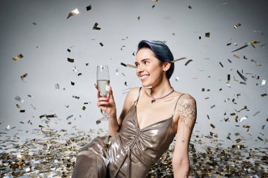A pretty young woman with short blue hair sits on the ground, elegantly holding a glass of champagne in a silver party dress.
