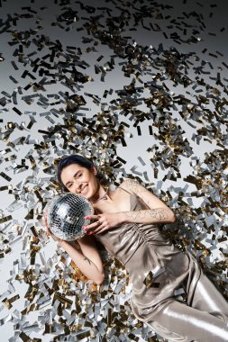 A pretty young woman with short blue hair lays on the ground, gracefully holding a shimmering disco ball.