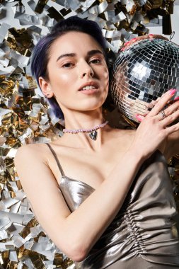 Young woman with blue hair poses elegantly in a silver dress, holding a sparkling disco ball in a studio setting.
