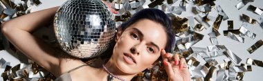 A pretty young woman with short blue hair holds a disco ball in front of her face, surrounded by a silver party dress.
